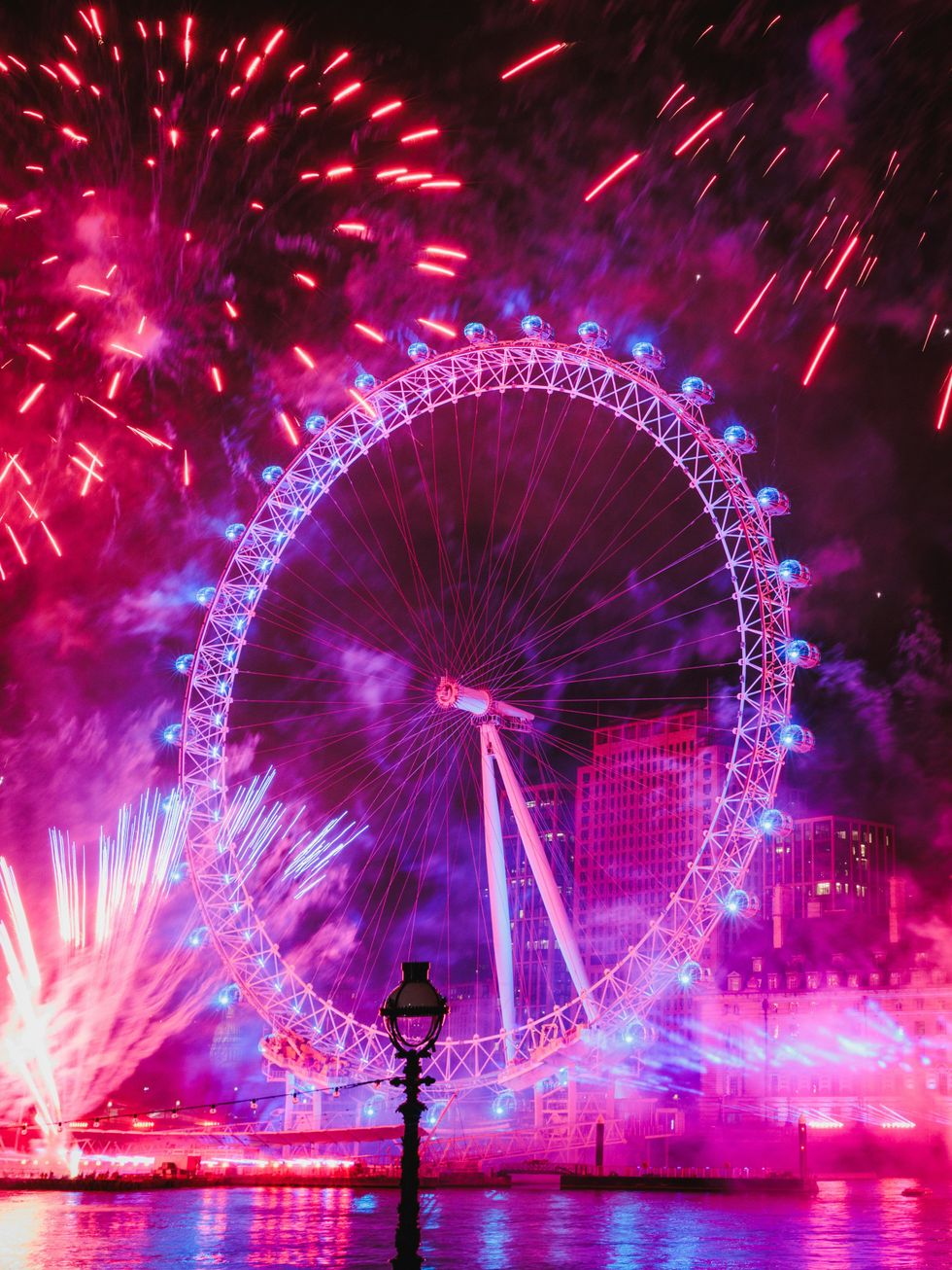 Fireworks explode around a lit Ferris wheel at night.