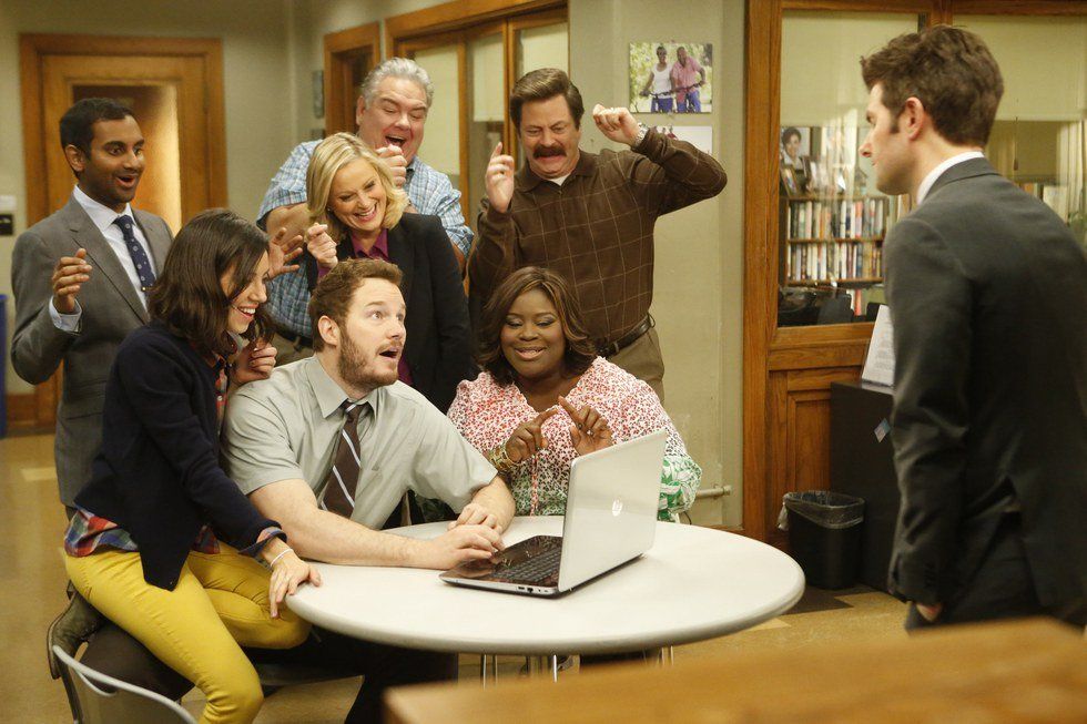 Parks and Recreation excitedly gathered around a laptop at a table in an office setting.